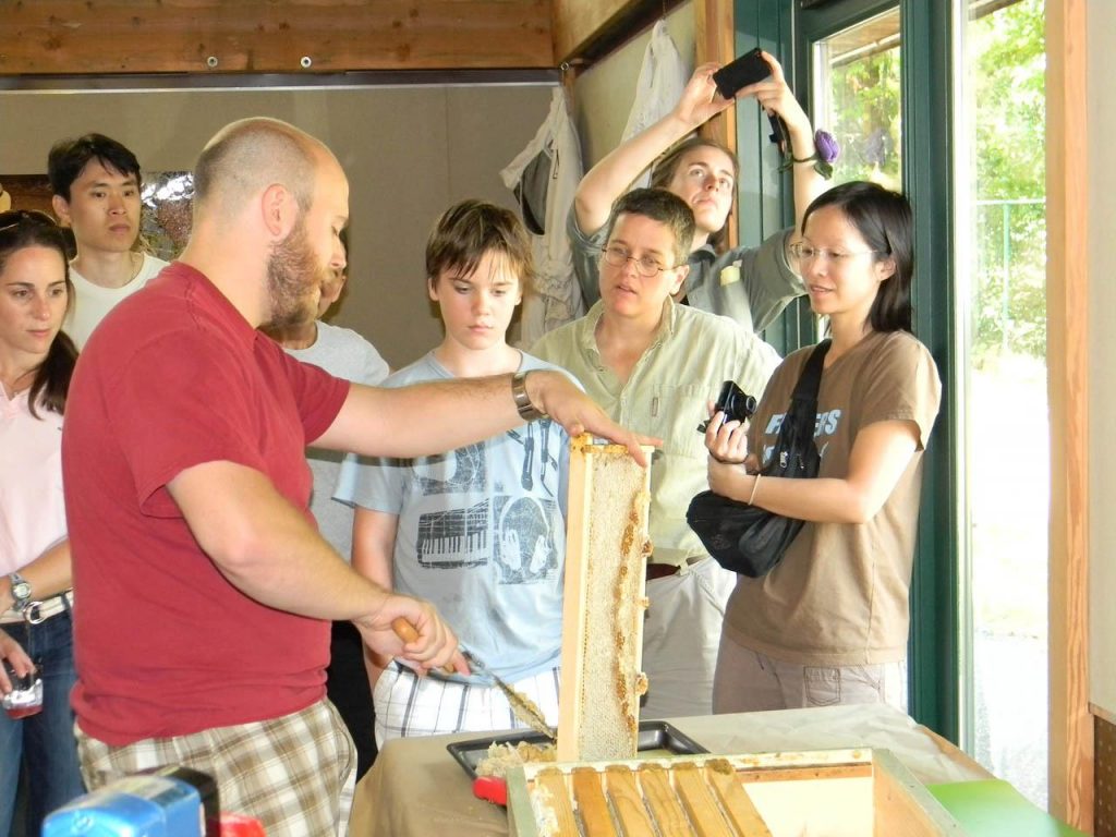 Matt Shoemaker holds a frame of honey and shows a group of people how to uncap the frame and extract honey from it